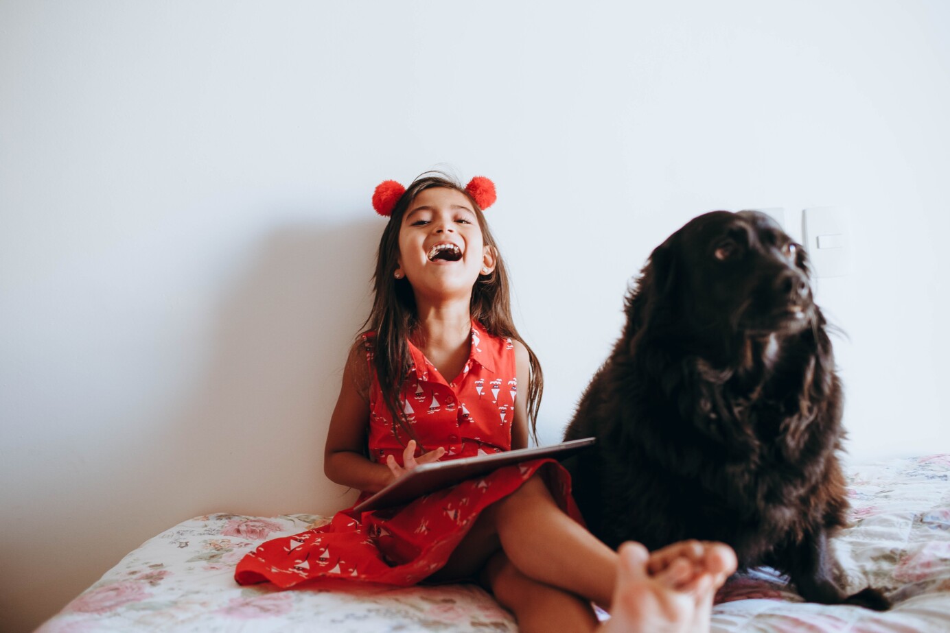 Lachend meisje in rood jurkje met een hond naast haar en een tablet op haar schoot